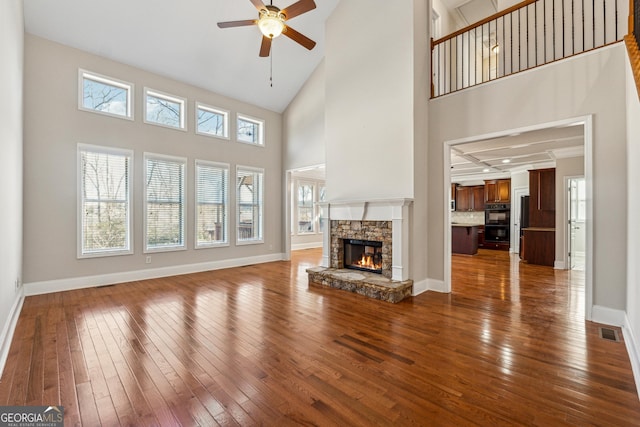 unfurnished living room with ceiling fan, a stone fireplace, hardwood / wood-style floors, and a towering ceiling