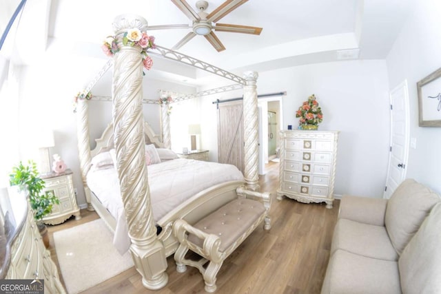 bedroom with ceiling fan, connected bathroom, wood-type flooring, a raised ceiling, and a barn door