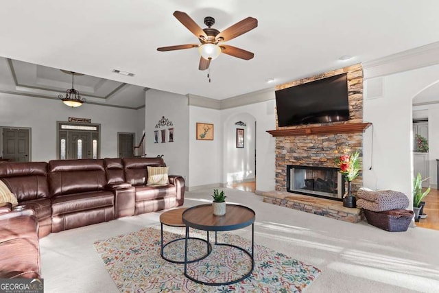 living room with a tray ceiling, a stone fireplace, crown molding, and ceiling fan