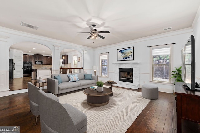 living room with crown molding, a stone fireplace, dark wood-type flooring, and ceiling fan