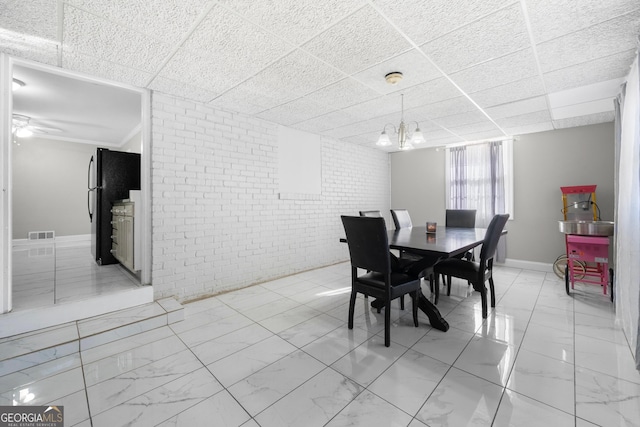 dining area featuring ceiling fan with notable chandelier, a paneled ceiling, and brick wall