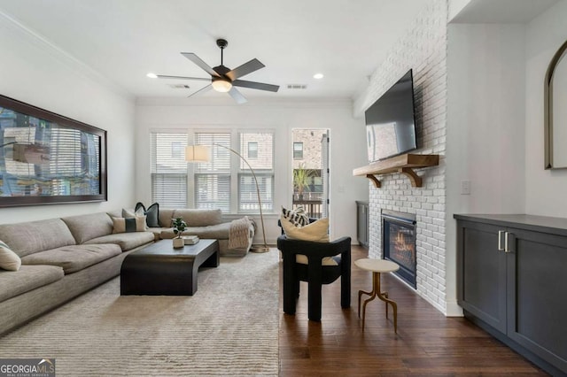 living room with dark wood-type flooring, ceiling fan, ornamental molding, and a brick fireplace