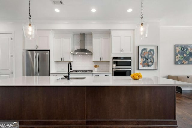 kitchen with wall chimney range hood, a large island with sink, pendant lighting, stainless steel appliances, and white cabinets