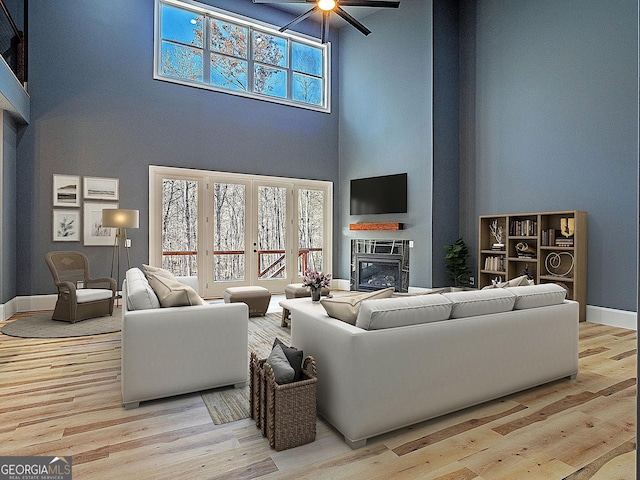 living room with light wood-type flooring, a glass covered fireplace, a towering ceiling, and a ceiling fan