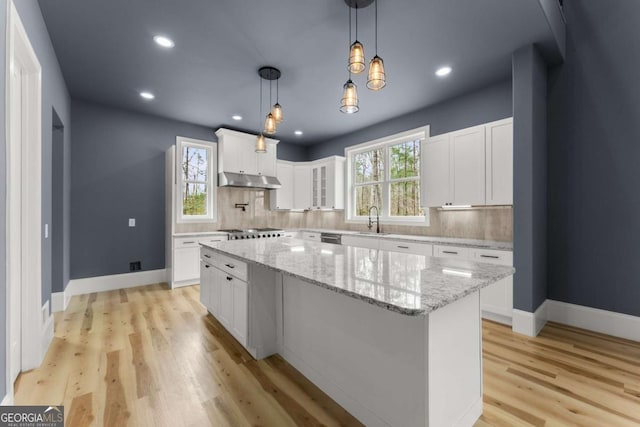 kitchen featuring white cabinets, a kitchen island, light stone counters, hanging light fixtures, and under cabinet range hood