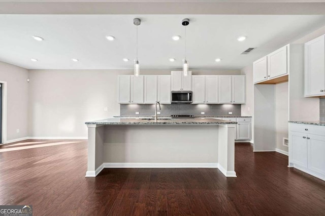 kitchen featuring dark wood finished floors, stainless steel microwave, a kitchen island with sink, a sink, and light stone countertops