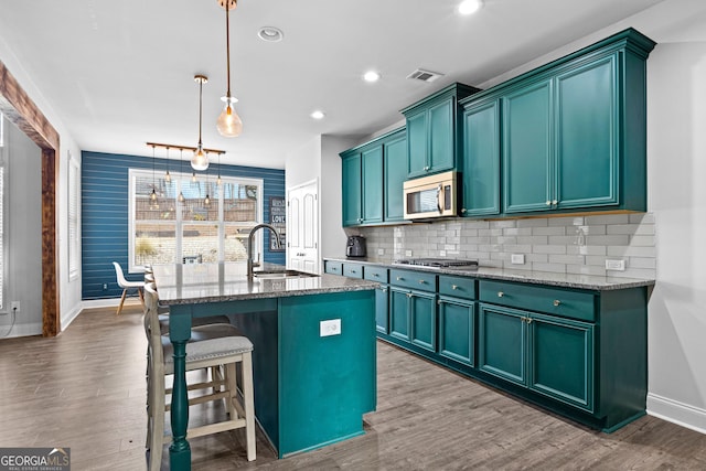 kitchen with sink, dark stone countertops, hanging light fixtures, stainless steel appliances, and an island with sink