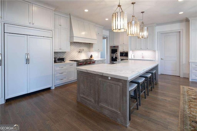 kitchen featuring appliances with stainless steel finishes, a breakfast bar, white cabinets, a large island with sink, and custom range hood