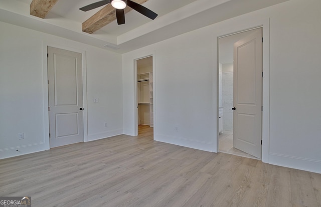 unfurnished bedroom featuring ensuite bathroom, a walk in closet, light hardwood / wood-style flooring, ceiling fan, and beam ceiling