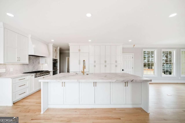 kitchen featuring a kitchen island with sink, white cabinetry, stainless steel appliances, light stone countertops, and custom exhaust hood