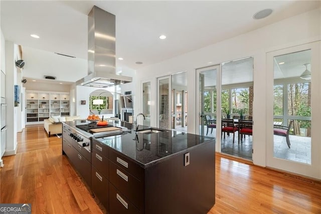 kitchen featuring dark brown cabinetry, light hardwood / wood-style floors, an island with sink, island exhaust hood, and dark stone counters