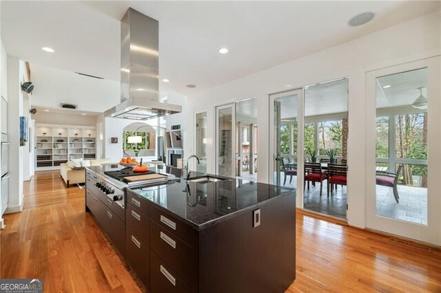 kitchen with dark brown cabinetry, light hardwood / wood-style flooring, an island with sink, island exhaust hood, and dark stone counters