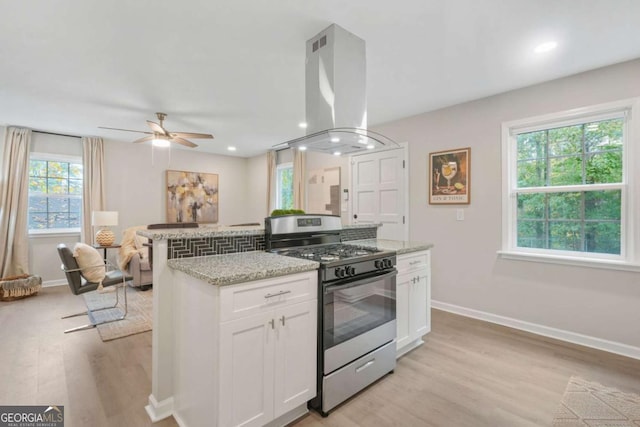 kitchen with white cabinets, island exhaust hood, light stone counters, stainless steel gas range, and light hardwood / wood-style flooring