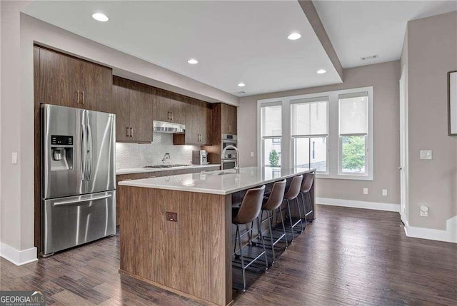 kitchen with an island with sink, backsplash, a kitchen bar, stainless steel appliances, and dark wood-type flooring