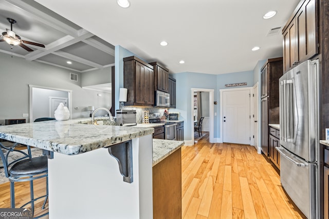 kitchen featuring coffered ceiling, a breakfast bar, light stone counters, appliances with stainless steel finishes, and kitchen peninsula