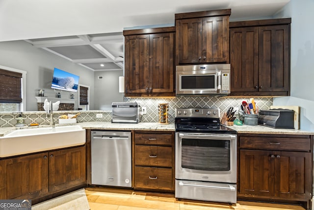 kitchen with sink, dark brown cabinets, stainless steel appliances, coffered ceiling, and light stone counters