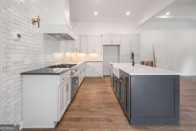 kitchen featuring dark wood-type flooring, custom exhaust hood, white cabinetry, a center island with sink, and stainless steel appliances