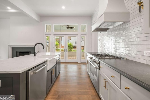 kitchen with sink, a kitchen island with sink, stainless steel appliances, decorative backsplash, and custom exhaust hood