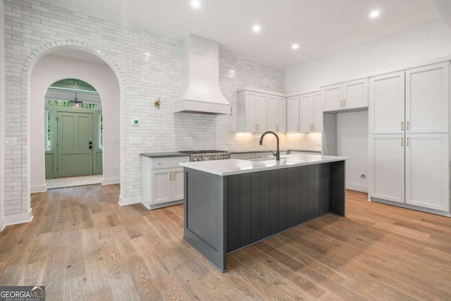 kitchen featuring sink, premium range hood, a kitchen island with sink, light hardwood / wood-style floors, and white cabinets