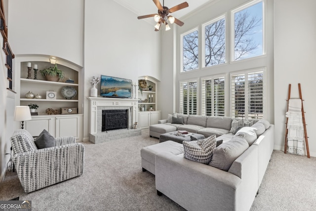 living room featuring built in shelves, crown molding, a brick fireplace, carpet flooring, and ceiling fan