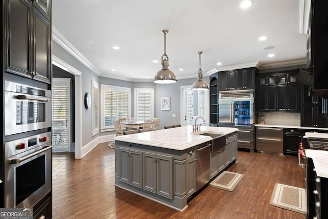 kitchen with pendant lighting, dark wood-type flooring, appliances with stainless steel finishes, light stone countertops, and a center island with sink