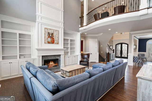 living room featuring crown molding, a towering ceiling, dark hardwood / wood-style floors, a tiled fireplace, and built in shelves