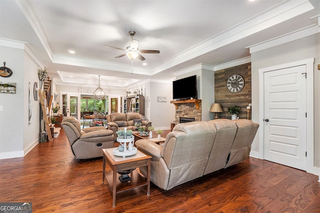 living room with ceiling fan with notable chandelier, a fireplace, a tray ceiling, crown molding, and dark wood-type flooring