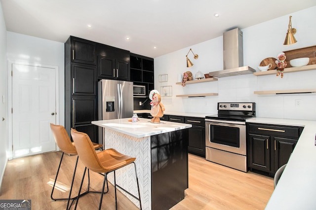 kitchen with appliances with stainless steel finishes, a kitchen bar, a center island, wall chimney range hood, and light wood-type flooring