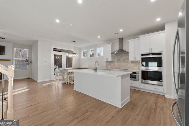 kitchen featuring a kitchen island with sink, wall chimney range hood, white cabinets, and appliances with stainless steel finishes