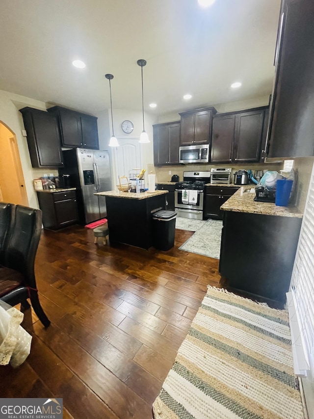 kitchen with dark wood-type flooring, hanging light fixtures, a kitchen island, stainless steel appliances, and light stone countertops