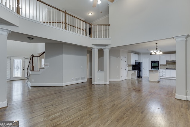 unfurnished living room featuring a towering ceiling, wood-type flooring, decorative columns, and ceiling fan with notable chandelier