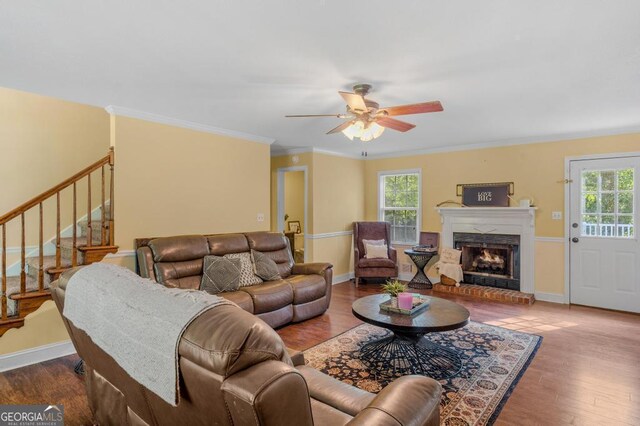 living room featuring a brick fireplace, wood-type flooring, ornamental molding, and ceiling fan