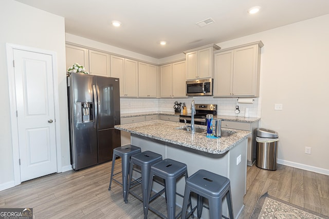 kitchen with light stone counters, light hardwood / wood-style flooring, stainless steel appliances, a kitchen island with sink, and decorative backsplash