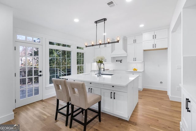 kitchen featuring sink, custom exhaust hood, hanging light fixtures, a center island with sink, and white cabinets