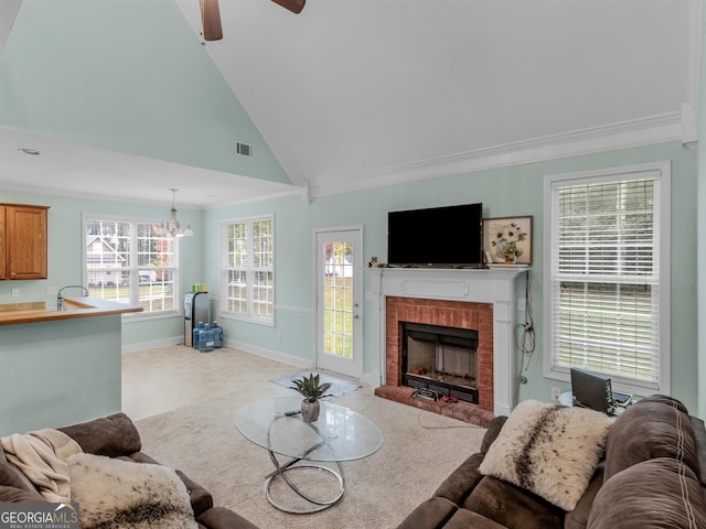 living room with ceiling fan with notable chandelier, high vaulted ceiling, light colored carpet, crown molding, and a brick fireplace