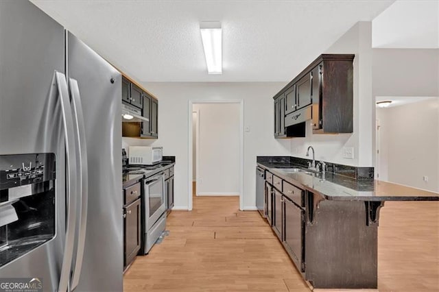 kitchen with dark stone counters, light wood-style flooring, range hood, stainless steel appliances, and a sink