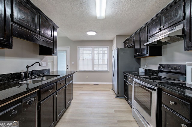 kitchen featuring light wood finished floors, stainless steel appliances, a sink, dark stone countertops, and under cabinet range hood