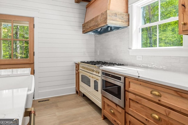 kitchen featuring light hardwood / wood-style flooring, appliances with stainless steel finishes, premium range hood, wooden walls, and decorative backsplash