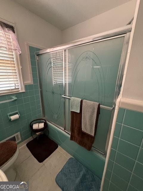 bathroom with tile walls, tile patterned flooring, combined bath / shower with glass door, and toilet