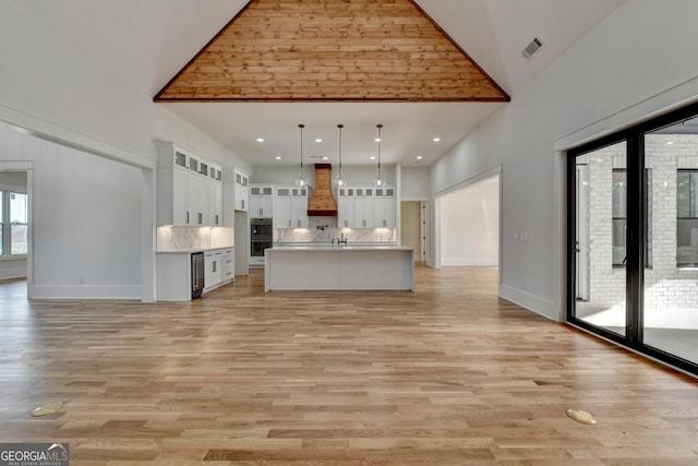 kitchen with premium range hood, pendant lighting, high vaulted ceiling, white cabinetry, and a kitchen island with sink