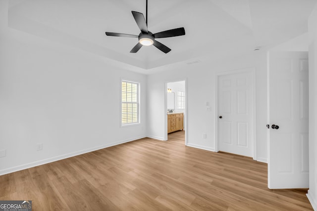 unfurnished bedroom featuring ceiling fan, ensuite bath, a raised ceiling, and light wood-type flooring