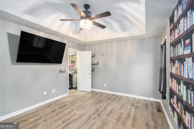 unfurnished bedroom with ceiling fan, a textured ceiling, a walk in closet, a raised ceiling, and light wood-type flooring