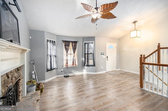 unfurnished living room with ceiling fan, a textured ceiling, a stone fireplace, vaulted ceiling, and light wood-type flooring