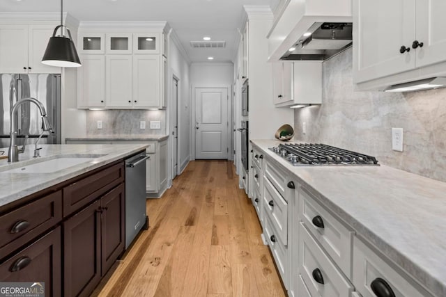 kitchen featuring wall chimney range hood, sink, white cabinetry, stainless steel appliances, and decorative light fixtures