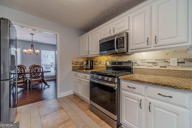 kitchen with white cabinets, appliances with stainless steel finishes, a notable chandelier, dark stone counters, and backsplash