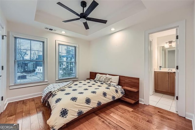 bedroom with a raised ceiling, connected bathroom, hardwood / wood-style floors, and ceiling fan