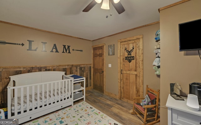 bedroom featuring crown molding, wood-type flooring, wooden walls, ceiling fan, and a crib