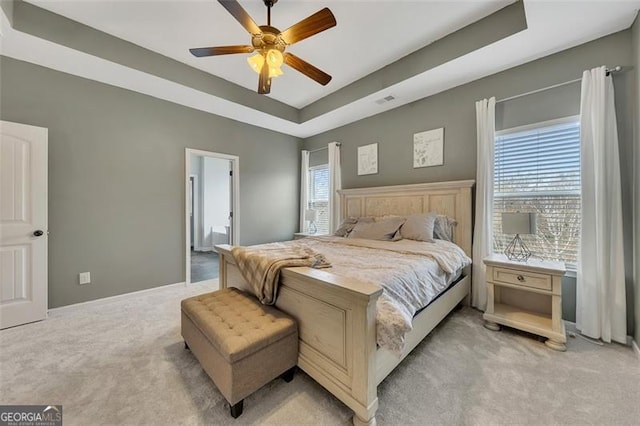 bedroom with ceiling fan, light colored carpet, a tray ceiling, and ensuite bath