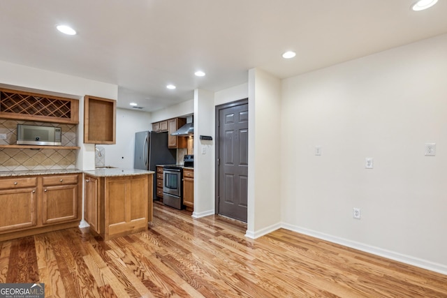 kitchen featuring wall chimney range hood, appliances with stainless steel finishes, light stone countertops, decorative backsplash, and light wood-type flooring