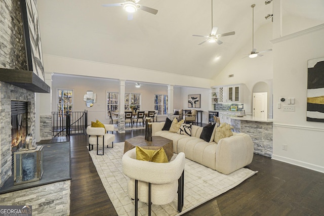 living area with high vaulted ceiling, a ceiling fan, a stone fireplace, and dark wood-style floors